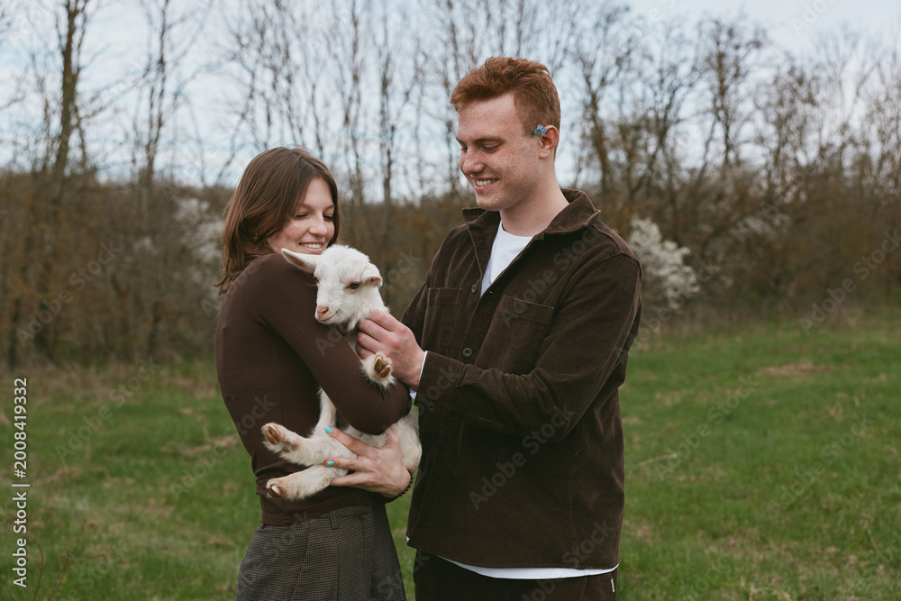 Fototapeta premium A happy couple outdoors with a small white goat kid, smiling as they cradle the playful animal in a green field, showcasing rural lifestyle and gentle interaction between humans and livestock.