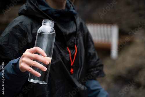 Woman hiker in raincoat holding water bottle outdoors in rainy weather during hiking, close-up view with copy space.