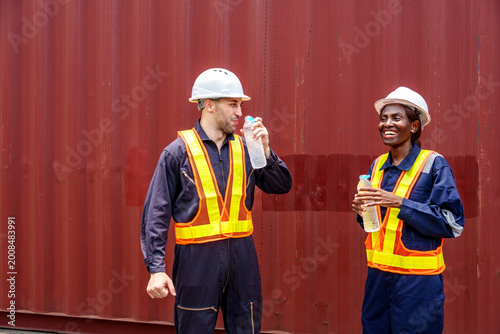 Smiling mid-adult Caucasian male and Black female industrial workers sharing a laugh while drinking water at an Asian container yard.