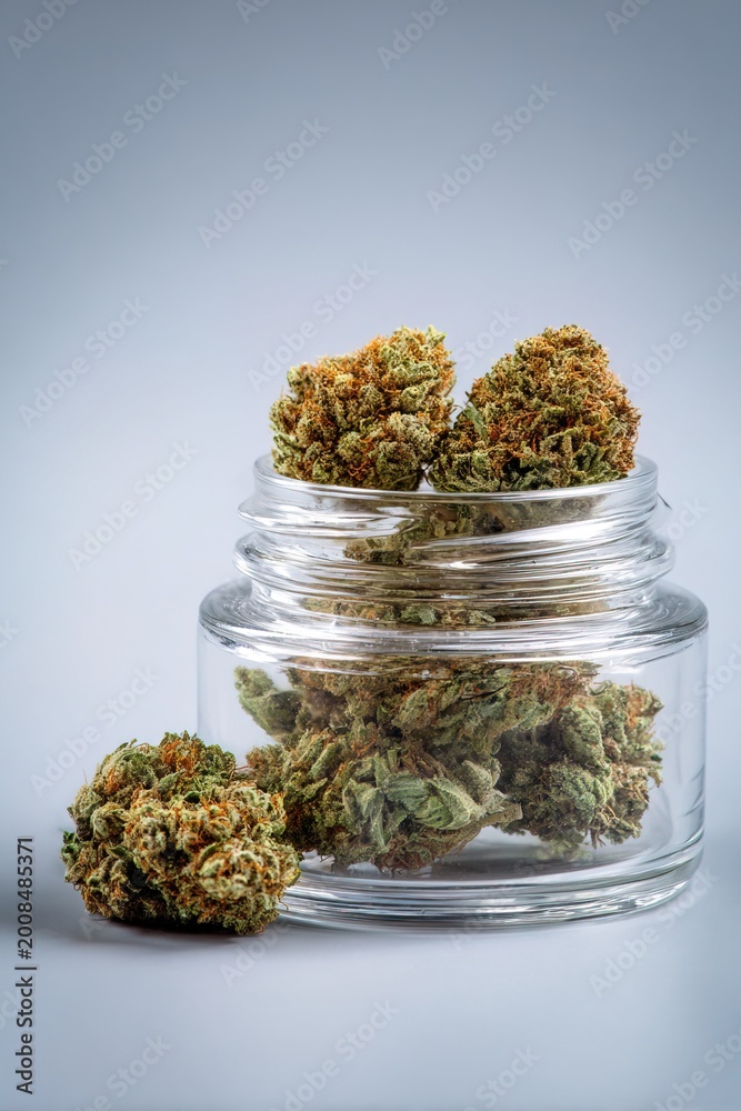 Obraz premium Buds of medical marijuana sit in a clear glass container with additional buds on a surface. The scene is well lit, highlighting the details of the buds