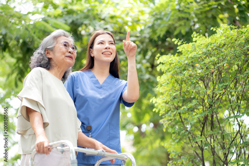 Asian female caregiver assistance senior patient with walker for physical rehabilitation and therapy, nurse supporting elderly woman walking in park, wellness and healthy retirement, medical concept.