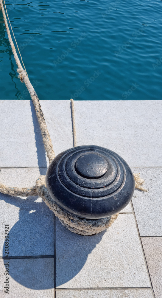Fototapeta premium A thick nautical rope is tied around a black cast iron mooring bollard on a stone pier in Baska on the island of Krk with blue sea water in the background