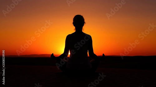 Silhouette of a young woman meditating in the lotus position on sand dunes during a vibrant orange sunset representing peace and mindfulness with a calm and serene mood