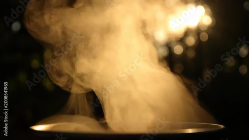 Thick steam billows dramatically from a circular surface while illuminated by a bright light source in the background creating a mystical and atmospheric effect