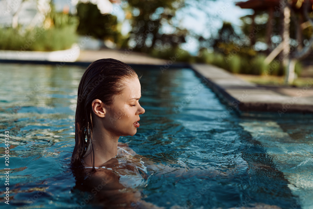 Fototapeta premium Young woman with wet hair relaxes in outdoor swimming pool during sunny day surrounded by trees and clear water lifestyle concept