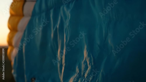 Close up of a blue and yellow paraglider wing fabric glowing in the warm sunset light while preparing for flight with a vast field in the background