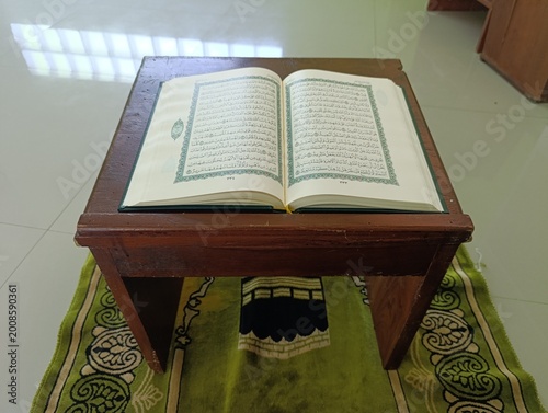 Open holy Quran on a wooden stand over a green prayer rug in indoor setting