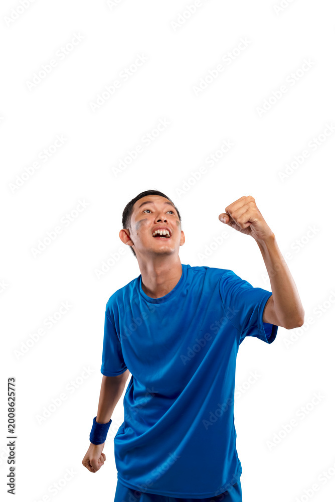 Naklejka premium Enthusiastic young Indonesian southeast asian man wearing a blue sports t-shirt and wristband, looking up and cheering with a clenched fist, celebrating a win or success against a white background