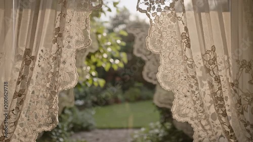 Delicate lace curtains gently billow in a sunlit window, revealing a lush garden view