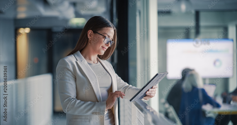 Obraz premium Businesswoman Uses a Digital Tablet in a Modern Office Hallway to Review Project Dashboards, Update Tasks, and Track Performance While a Team Collaborates in a Glass Meeting Room
