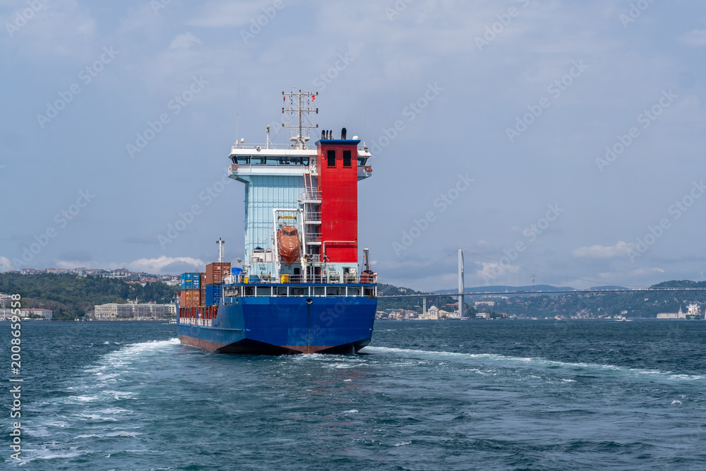 Fototapeta premium A cargo ship moves through the Bosphorus Strait, leaving a wake behind as it approaches the urban scenery, showcasing maritime activity on a sunny day.