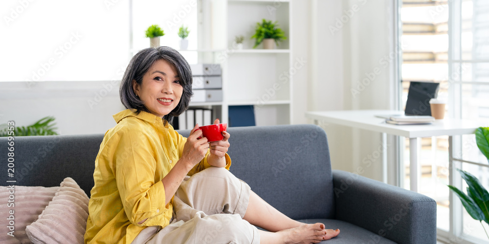 © Fahng - Happy relaxed middle aged asian woman drinking coffee relaxing on sofa at home. Smiling stylish middle aged 50s lady enjoying resting sitting on couch.