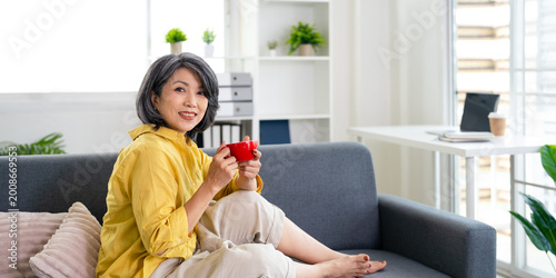 Happy relaxed middle aged asian woman drinking coffee relaxing on sofa at home. Smiling stylish middle aged 50s lady enjoying resting sitting on couch.