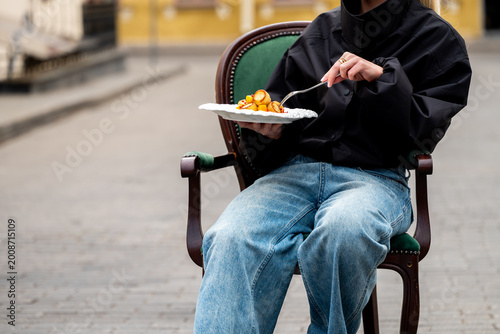 Slika na platnu A person in a black jacket and blue jeans sits in a green vintage chair on a city street, holding a white plate of food and a fork
