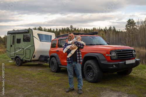 A man holds a baby while standing next to an orange off-road vehicle and a travel trailer