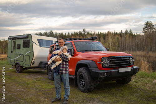 A smiling father holds his baby in front of an orange car and a travel trailer