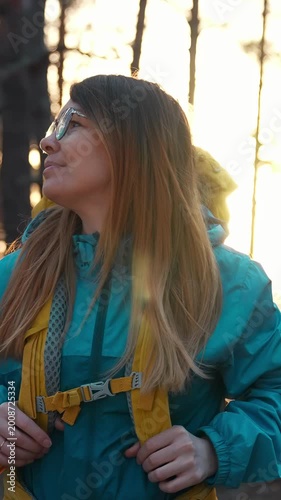 A woman stands outdoors, looking up, with sunlight filtering through the trees behind her. She wears a teal jacket and has long brown hair, embodying a spirit of adventure in nature.