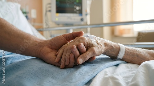 A person comforts an elderly patient in a hospital bed holding hands