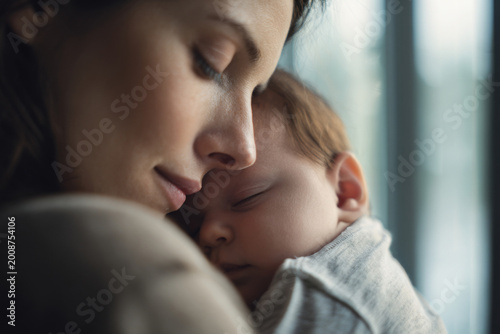 Mother holding sleeping baby near large window with gentle daylight, medium close-up composition, calm peaceful mood, soft background blur