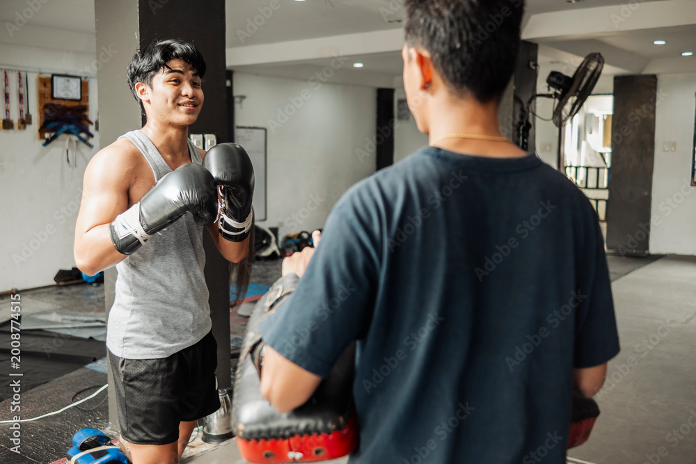 Fototapeta premium Young Asian boxer playfully sparring with coach holding Muay Thai pads, preparing for striking drills in a gym.