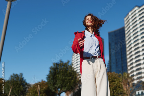 A young woman with curly hair stands in an urban outdoor setting, wearing a red jacket over a white shirt and beige pants, smiling confidently as the city skyline rises behind her.