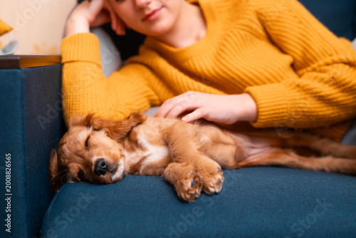 A young woman with brown hair sits on a sofa, gently petting a golden retriever puppy that is sleeping beside her