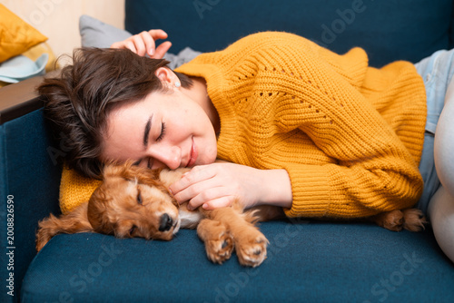 A young woman lies on a couch, smiling as she cuddles with a sleeping puppy. The setting is indoors, creating a warm atmosphere during leisure time together