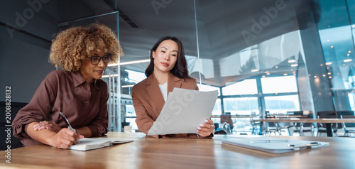 Obraz na płótnie Two professional women businesswomen in modern office reviewing document, one taking notes at table, business meeting, teamwork, coaching and analysing paperwork collaboration