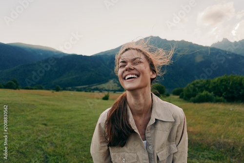 Happy woman enjoying outdoor life, smiling in a grassy field with mountains in the background, casual jacket, sunlit moment, lifestyle portrait of carefree everyday joy