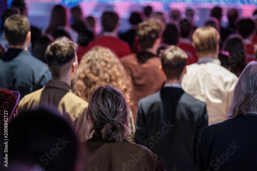 Back view of standing audience at indoor event, diverse people focused on stage, soft colorful lighting.
