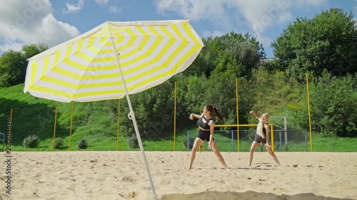 Two women perform yoga stretches on sandy beach, embracing warmth of sun. Vibrant umbrella stands nearby, adding color to this peaceful moment. Beach yoga. 4K, UHD