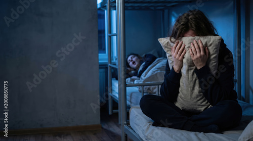 Woman covering ear with pillow because another woman is snoring in bedroom at night. Sleep problem and insomnia concept. Tired roommate suffering from loud snoring partner in dormitory.