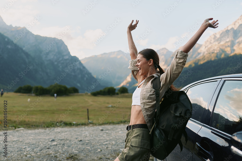 Obraz premium Smiling woman with a backpack beside a car in a mountain valley, embracing outdoor life, nature escape, and wellness. Bright, airy moment with adventurous spirit. Casual outdoor vibes with travel