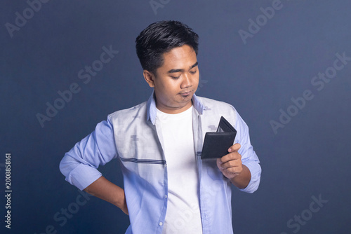 Photography Young man looking down at his open black wallet with a pouting and contemplative expression against a dark blue background