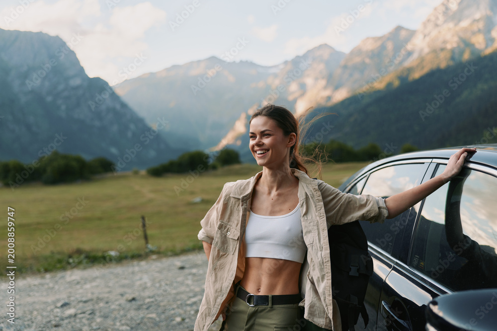 Fototapeta premium Smiling woman in casual clothes leaning on car door in mountain landscape. Outdoor lifestyle, travel, freedom, and adventure concept with natural light and relaxed mood.