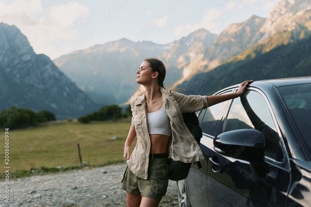 Fototapeta premium Young woman in casual outfit leaning on car outdoors with mountains in background. Concept of lifestyle, travel, freedom, and adventure in nature.