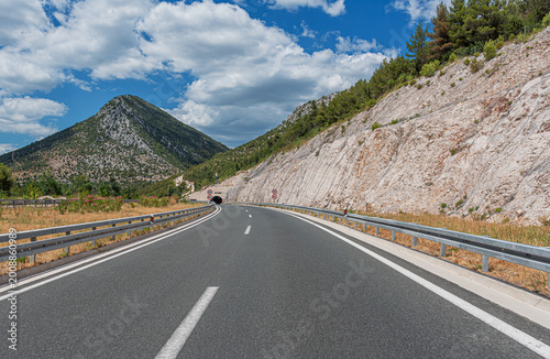 An empty asphalt highway curves through a mountainous landscape toward a tunnel under a bright blue sky with white clouds.