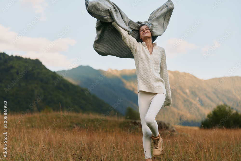 Fototapeta premium Woman dances in a field near mountains, wearing white sweater and pants, scarf billowing, carefree lifestyle shot capturing joy, movement and outdoor freedom