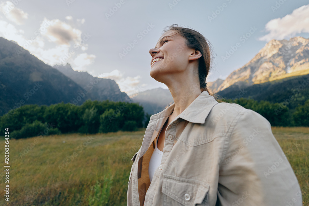Fototapeta premium Cheerful woman outdoors, smiling with eyes closed, sunlit moment in a grassy field, mountains in the background, relaxed posture, natural colors, lifestyle vibe