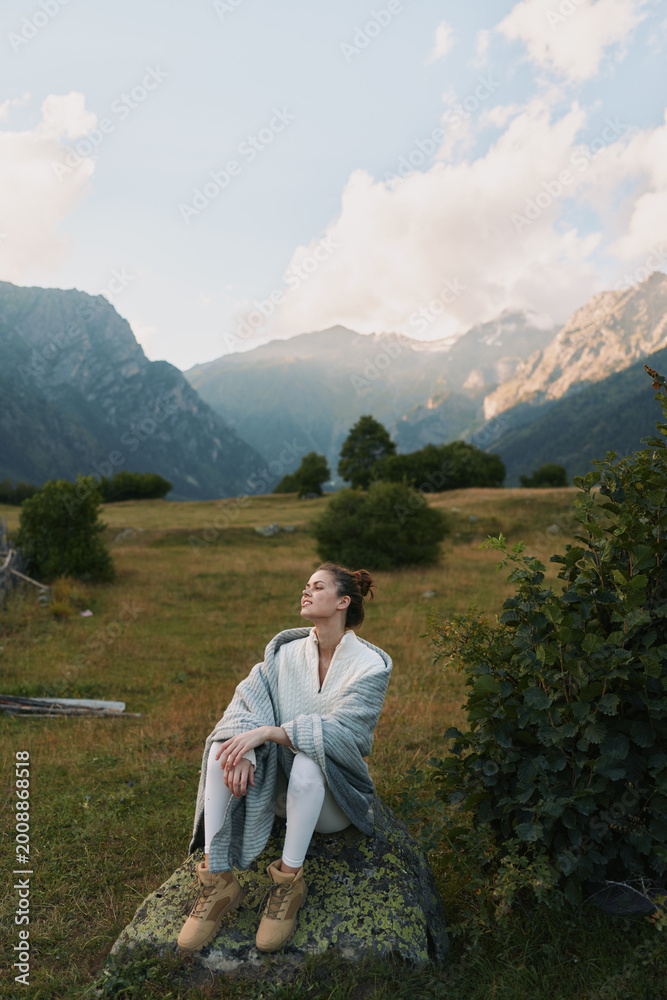 Fototapeta premium Woman in a cozy shawl sits on a rock in a mountain meadow, enjoying tranquil nature and a relaxed casual lifestyle portrait amid open sky and distant peaks