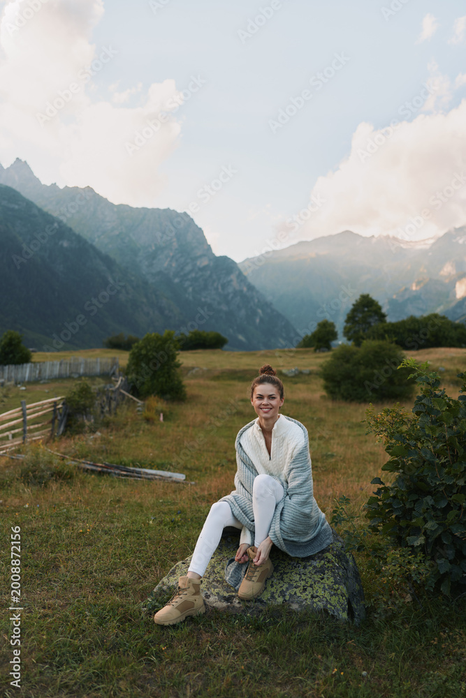 Fototapeta premium Casual joyful woman sits on a rock in a scenic mountain meadow, wearing a cardigan and white jeans, smiling, enjoying outdoor life in a peaceful rural landscape under soft daylight.