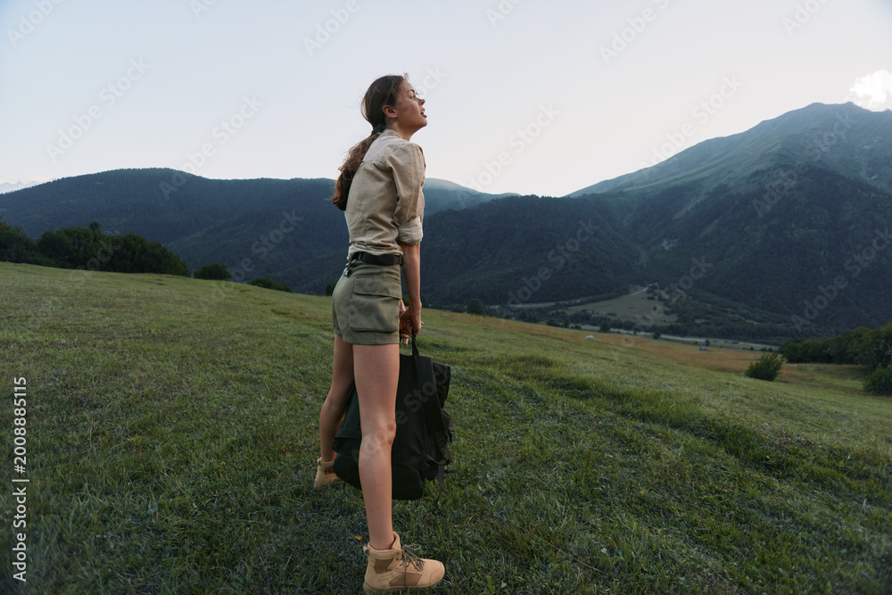 Fototapeta premium Young woman explores a meadow in the mountains wearing a beige shirt and olive shorts, carrying a dark tote, enjoying fresh air and open space under a clear sky