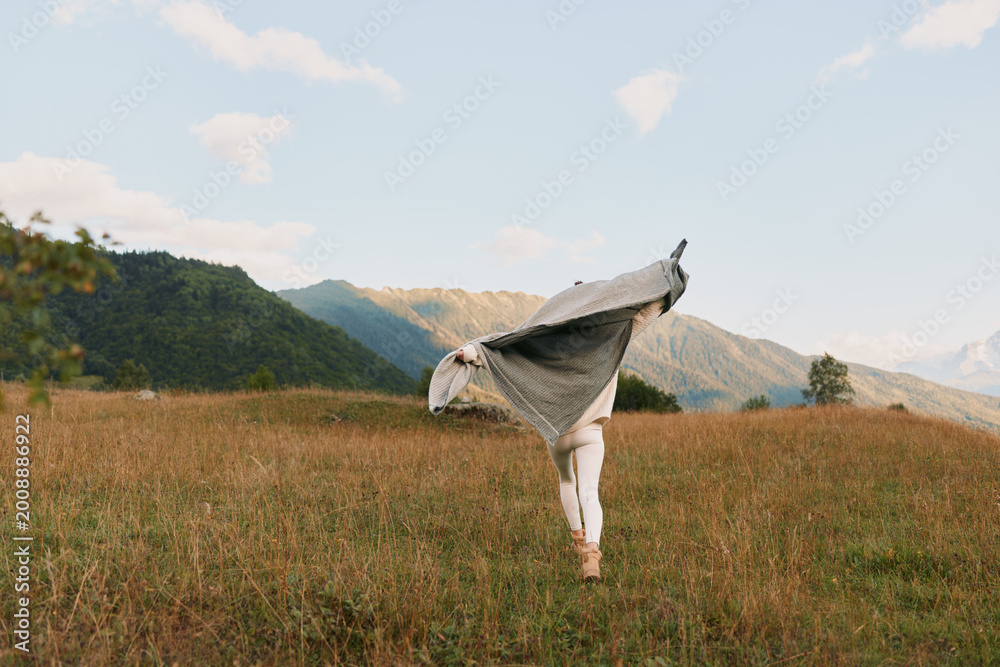Fototapeta premium A woman in a light shawl stretches in a golden meadow with distant mountains, conveying freedom and calm in the open countryside, an airy scene of movement under a bright sky.
