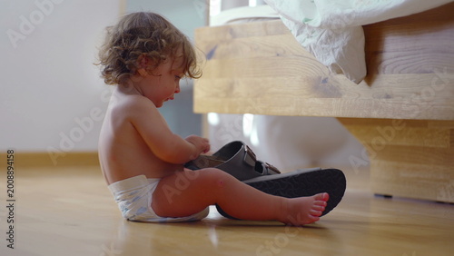Konstfotografi Toddler sitting on bedroom floor lifting sandal to examine sole closely, focused