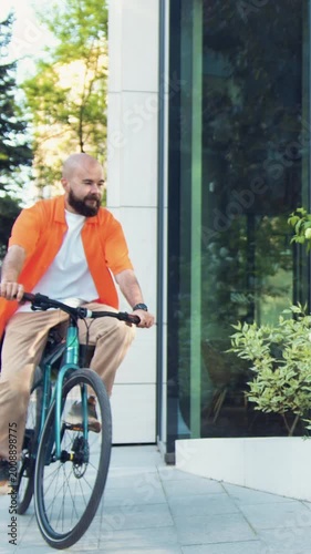 Handsome bearded smiling man riding bicycle at downtown district. Professional young male going to work and typing by phone while sitting on the bench with bicycle in background.