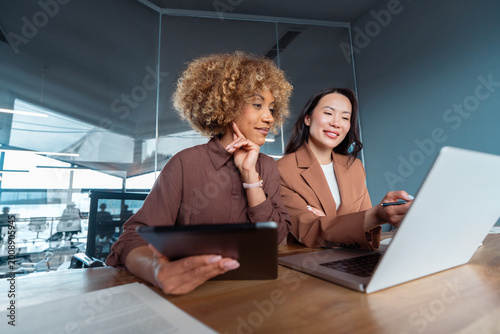 Two professional women collaborating at office desk, reviewing data on laptop and tablet in meeting room, teamwork and business planning. Team of women using technology at work, doing analysing