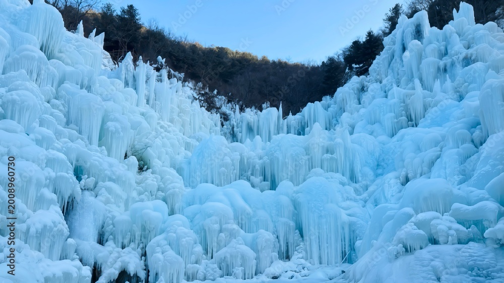 Obraz premium Stunning ice wall with giant icicles and frozen formations in a winter valley under a clear blue sky.