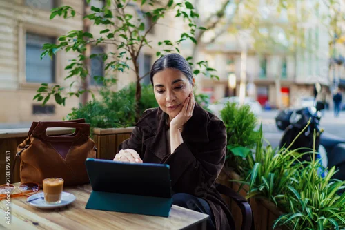 Obraz Thoughtful Hispanic woman working outdoors at cafe table with tablet, hand on face, reflecting focus, mindfulness, and urban freelance lifestyle surrounded by greenery.