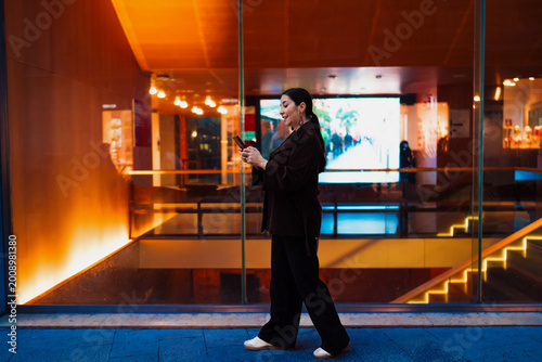 Obraz na plátne Hispanic woman walks past glass wall while focused on smartphone, representing on-the-go connectivity, tech fluency and urban independence in mobile professional life