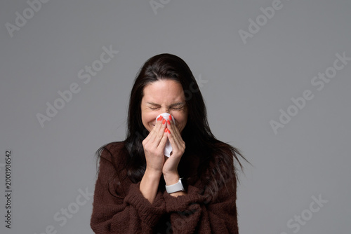Sick woman sneezing into white tissue holding napkin with both hands isolated on grey background